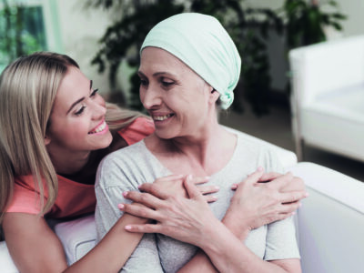 A woman with cancer is sitting on a white sofa next to her daughter. A girl is hugging a woman. They are sitting in the lobby of a modern clinic. Around them flowerpots with flowers.