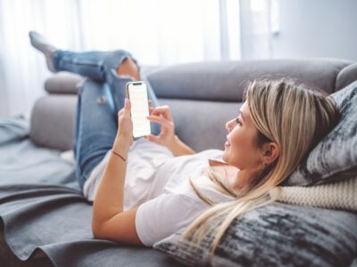 Charming caucasian blond young woman dressed casual lying down on sofa in living room, relaxing and using smart phone.